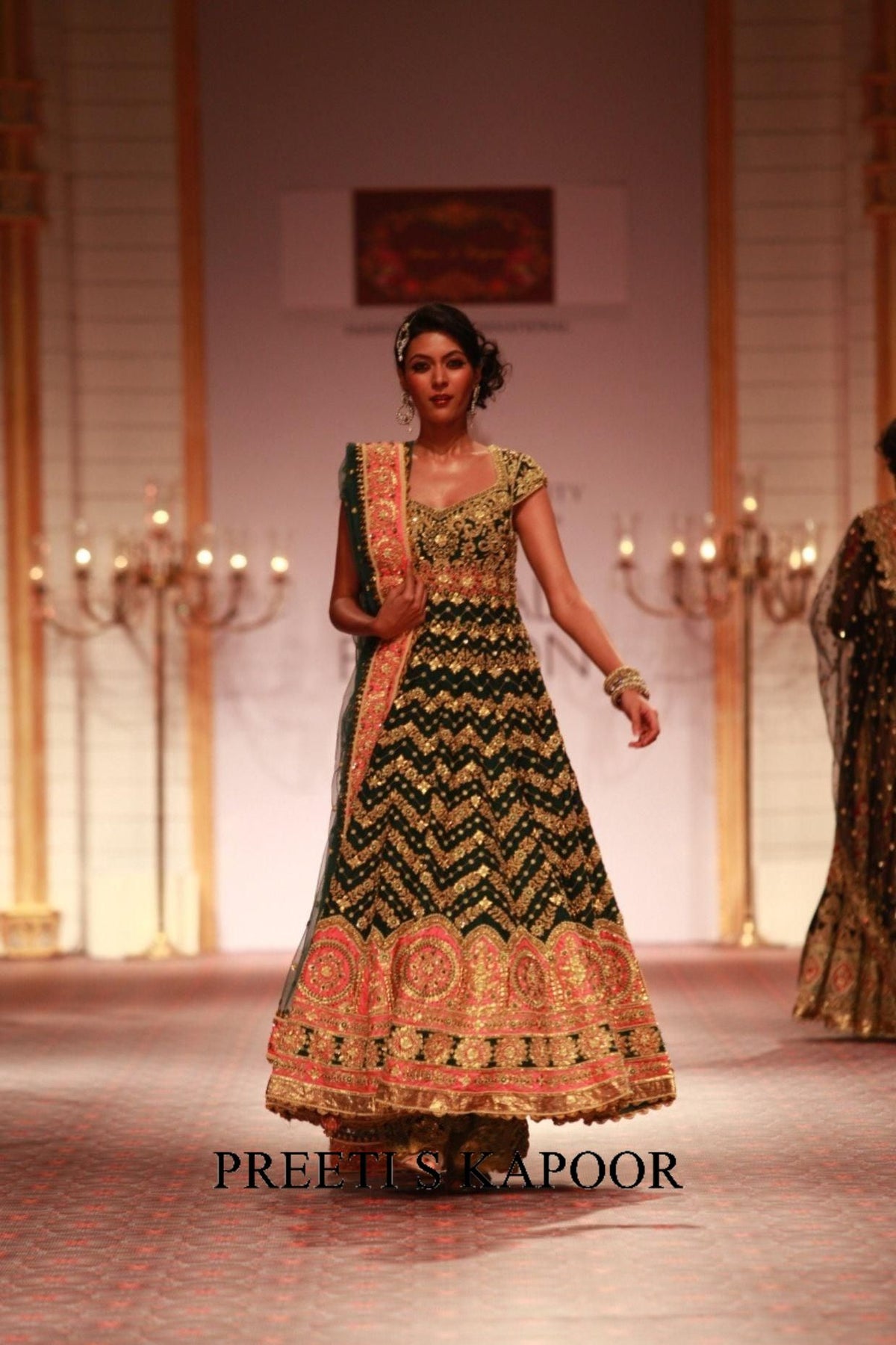 Model wearing a traditional embroidered dress on a runway with 'Preeti S Kapoor' branding.
