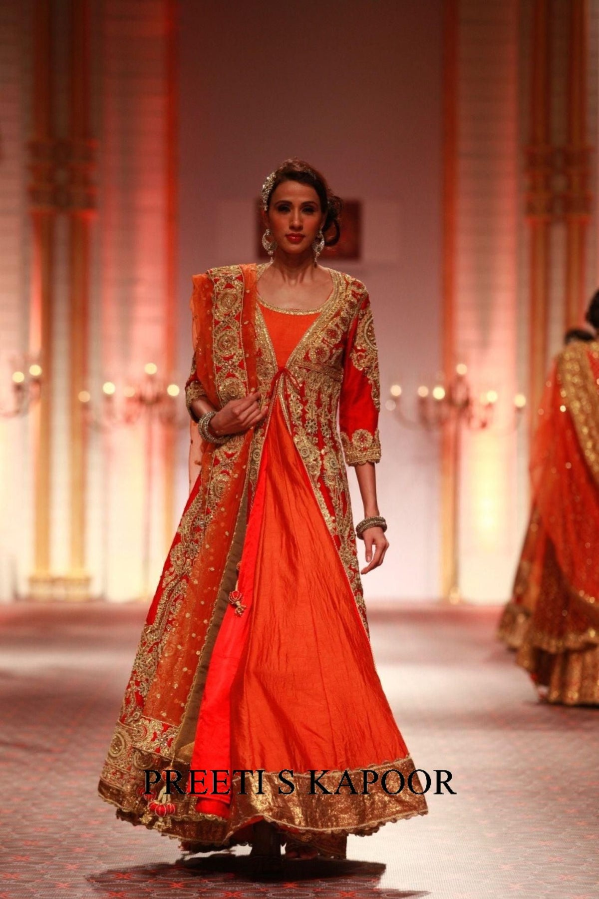 Model wearing a traditional orange and gold outfit on a runway with 'Preetis Kapoor' branding.