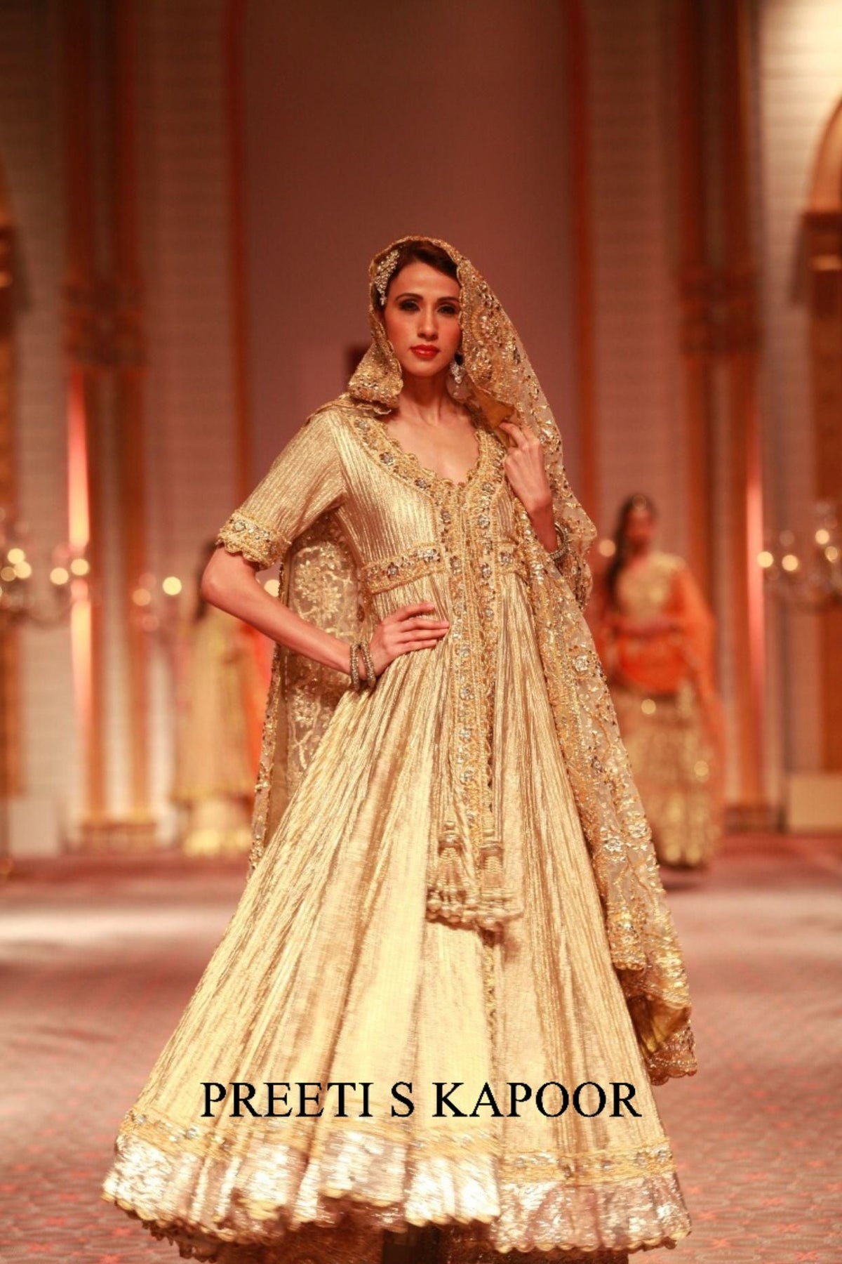 Model wearing a gold embroidered traditional outfit on a runway with 'Preeti S Kapoor' branding.