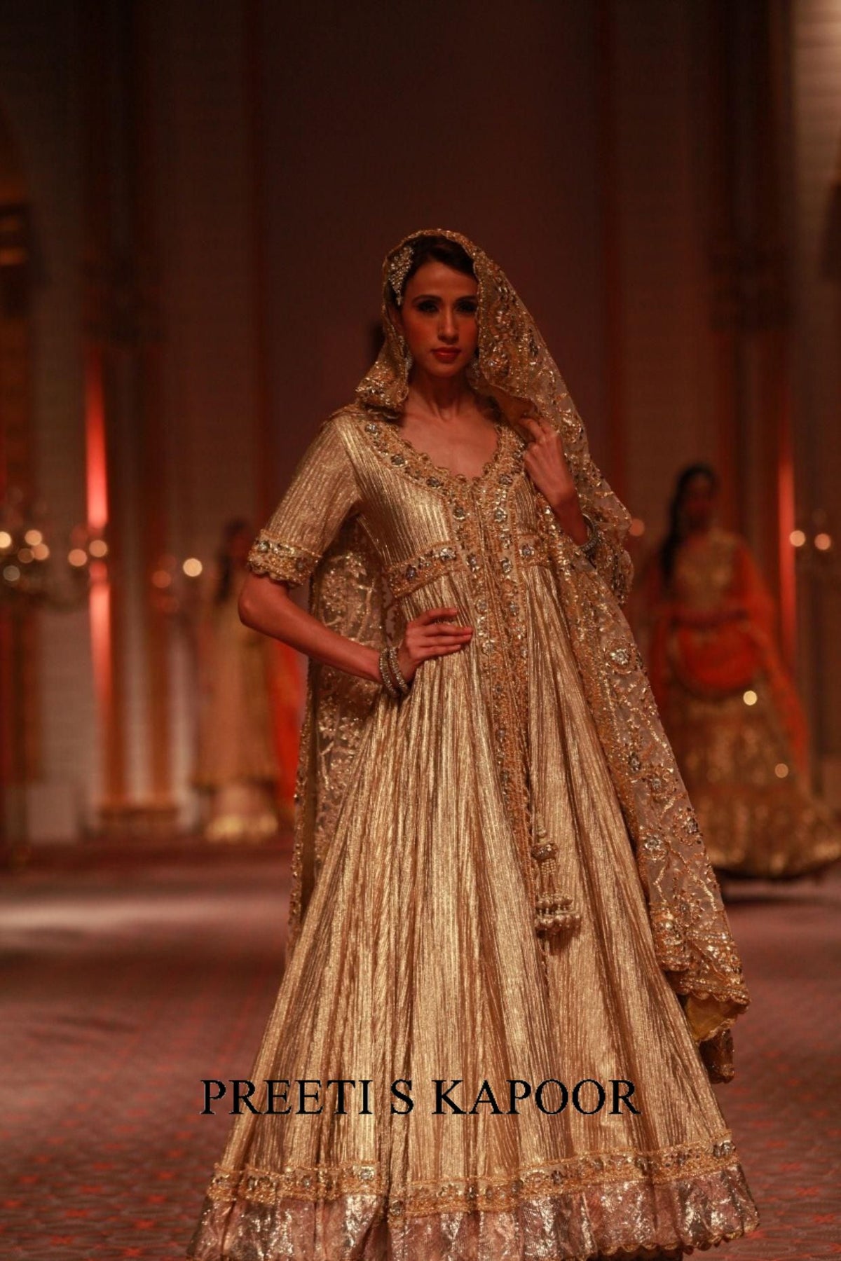 Woman in a traditional embroidered outfit with a veil at an event, wearing 'Preeti S Kapoor' brand.