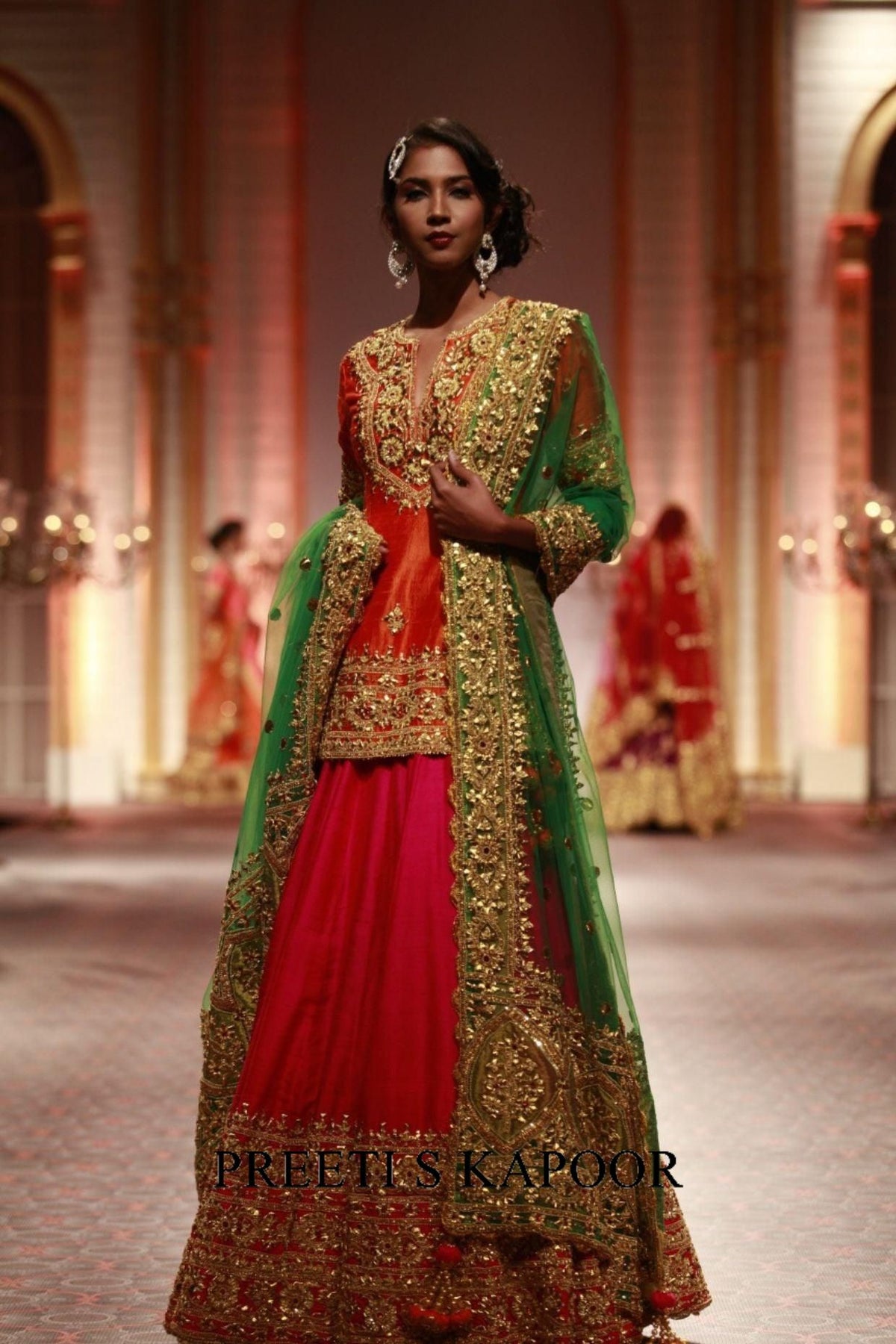 Model wearing a green and red embroidered traditional outfit on a runway with 'Preeti S Kapoor' branding.
