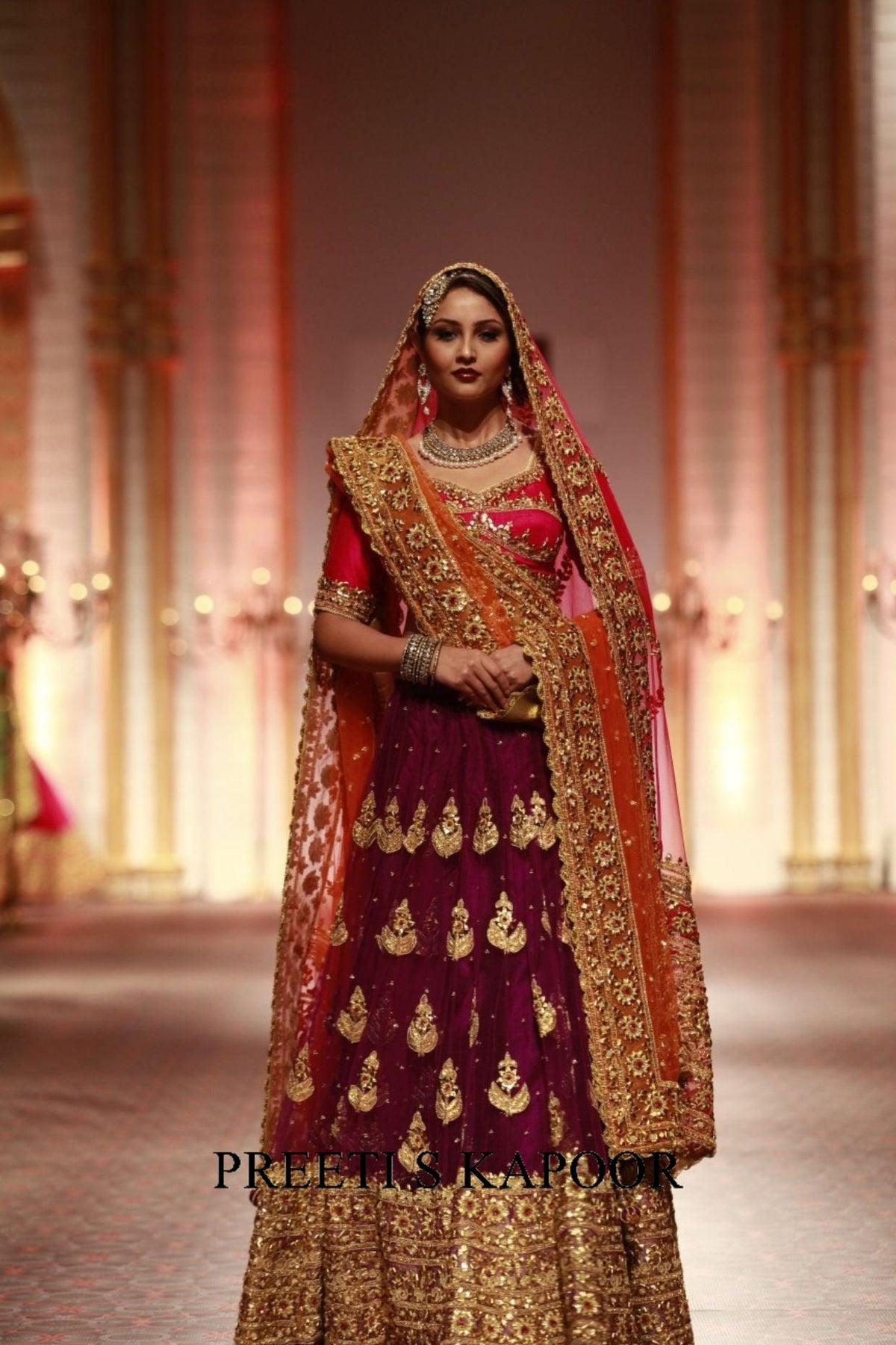 Woman in traditional Indian bridal attire with intricate designs and jewelry, standing on a stage.