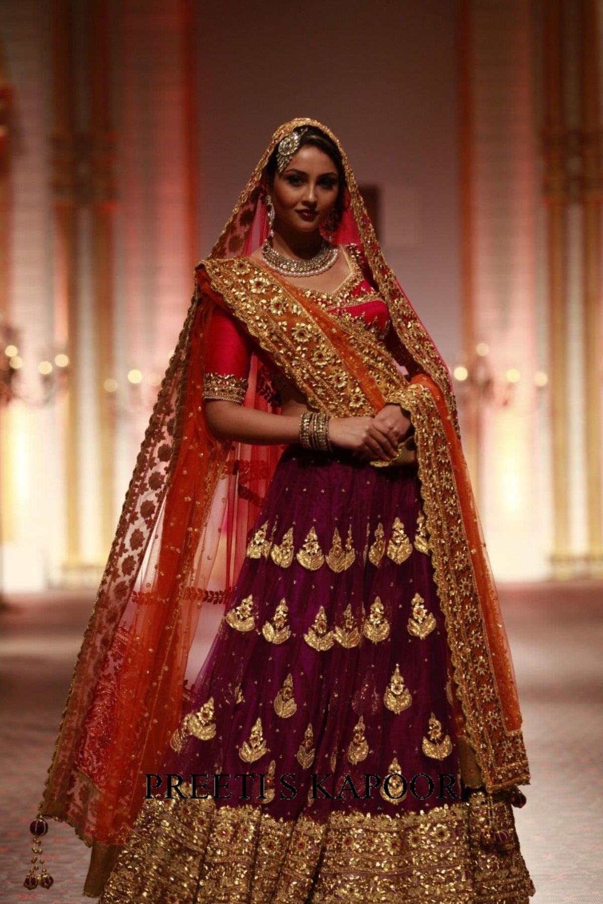 Woman in traditional maroon and gold embroidered outfit with a red dupatta on a blurred background