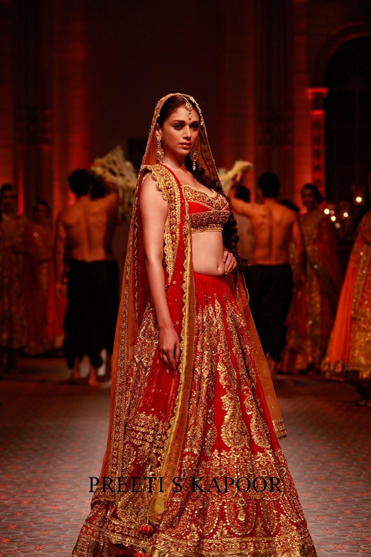 Model wearing a red and gold embroidered lehenga on a runway with 'Preeti S Kapoor' branding.