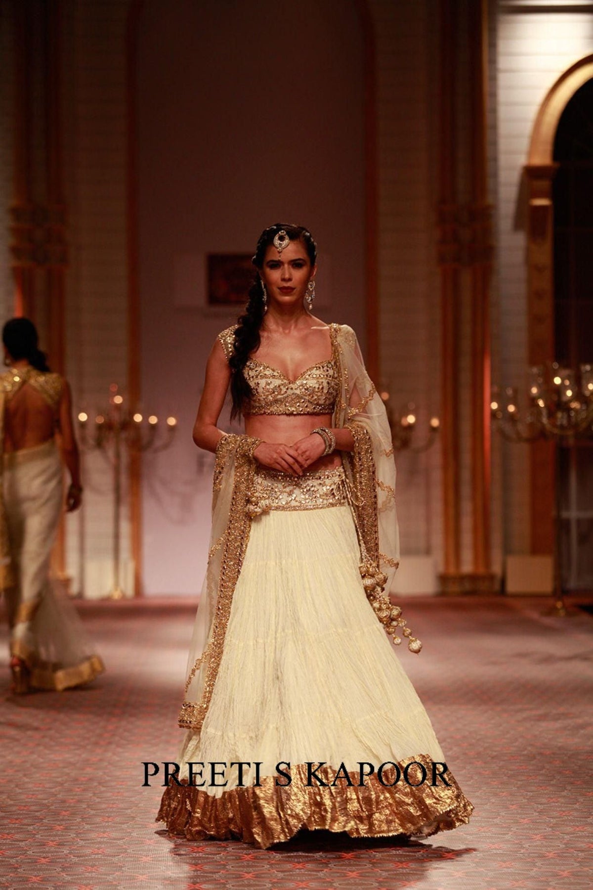 Model wearing a gold and white traditional outfit on a runway with 'Preeti S Kapoor' branding.