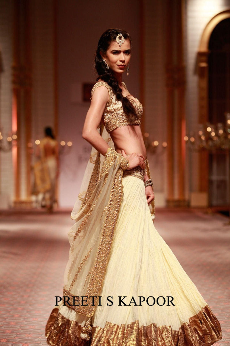 Model wearing a traditional outfit with gold and white design, standing on a stage with 'PREETI S KAPOOR' branding.
