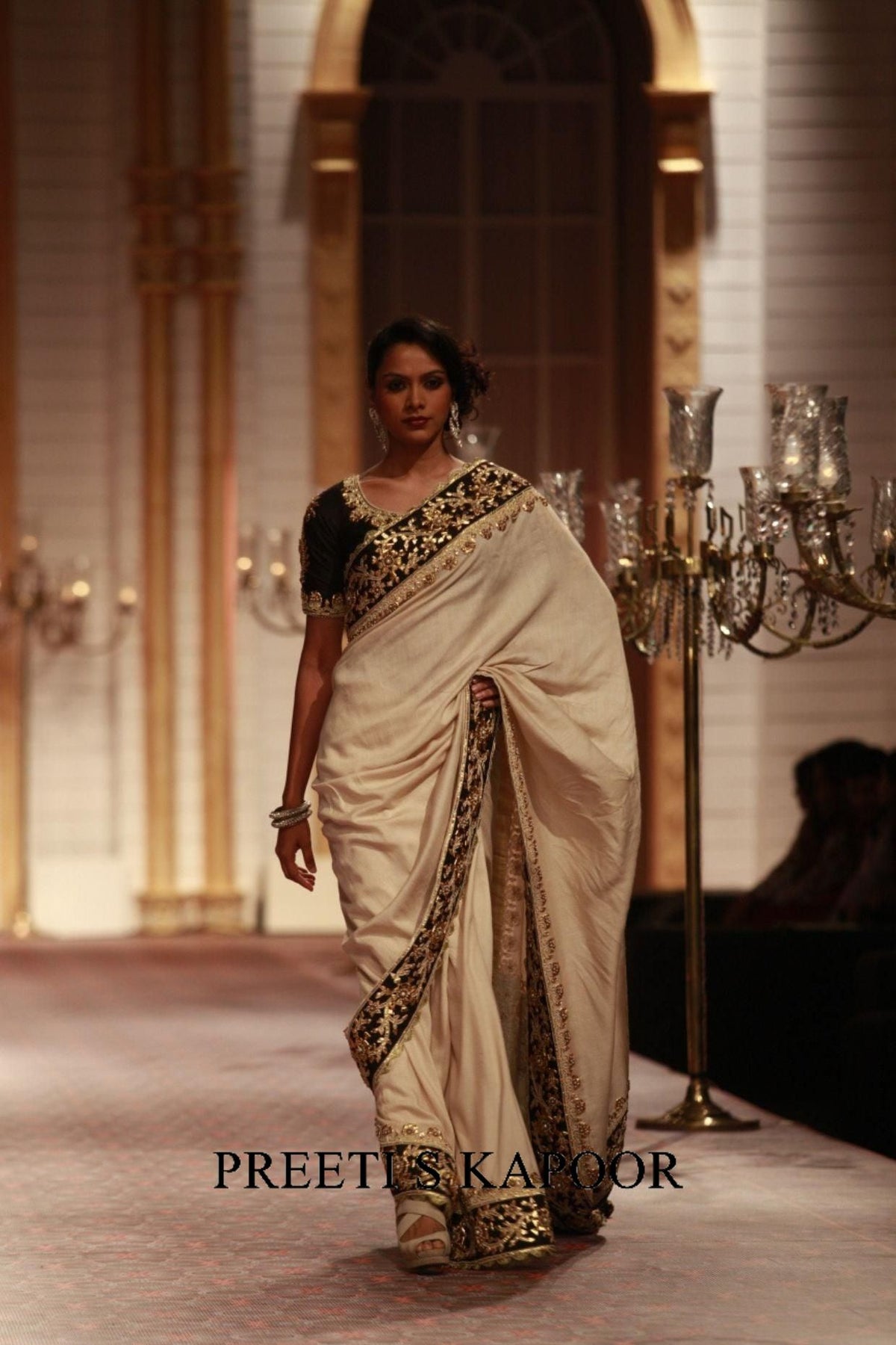Woman in a white saree with black and gold patterns on a runway, with 'Preetis Kapoor' branding.