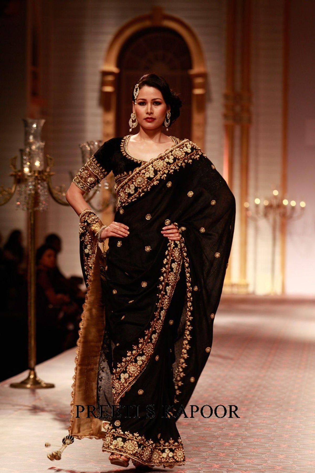 Woman in a black saree with gold embroidery on a stage, with 'Priti Lele Kapoor' branding.