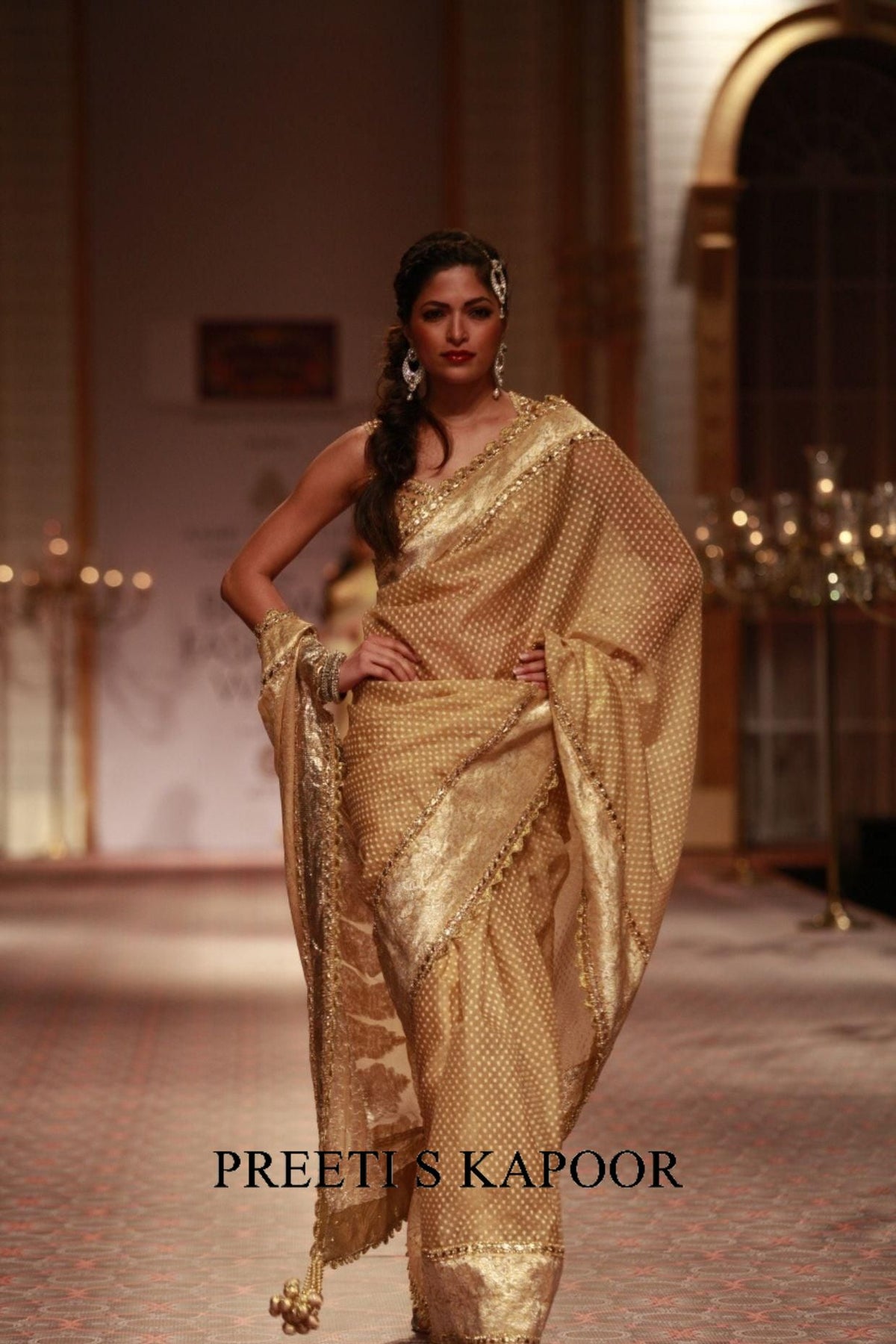 Woman in a gold saree with a blurred background, featuring the brand 'Preeti S Kapoor'.