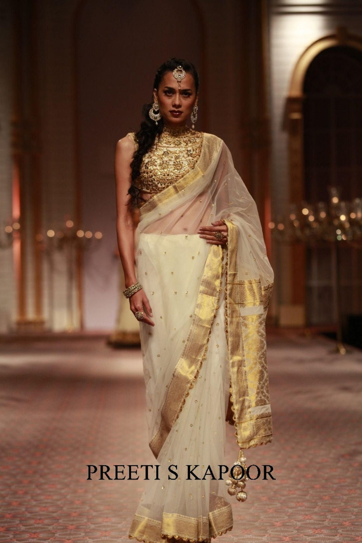 Woman in a traditional outfit with gold and white saree at an event, Preeti S Kapoor brand visible.