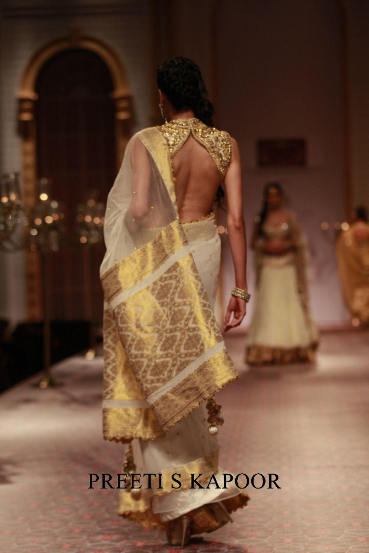 Model wearing a gold and white saree with a blurred background, featuring the brand 'Preeti S Kapoor'.