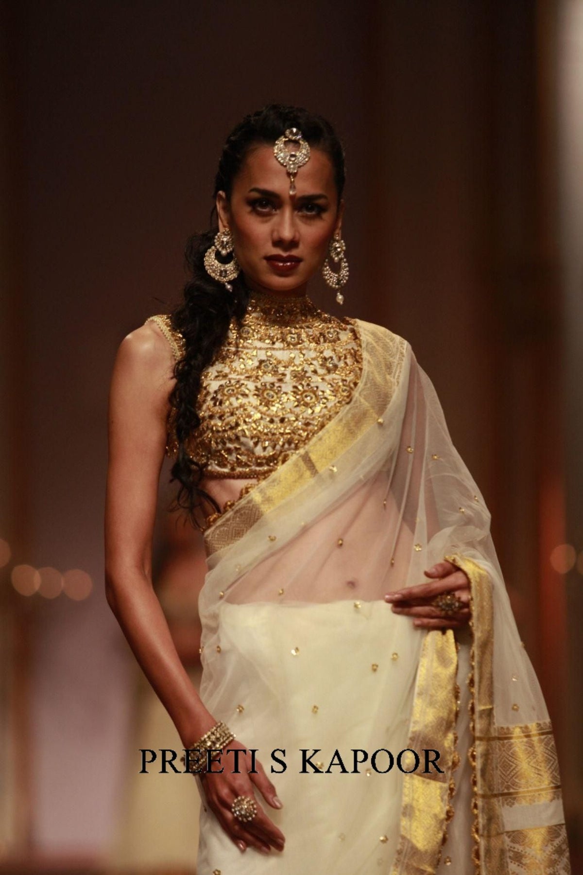 Model wearing a traditional outfit with gold and white elements, posing against a blurred background.