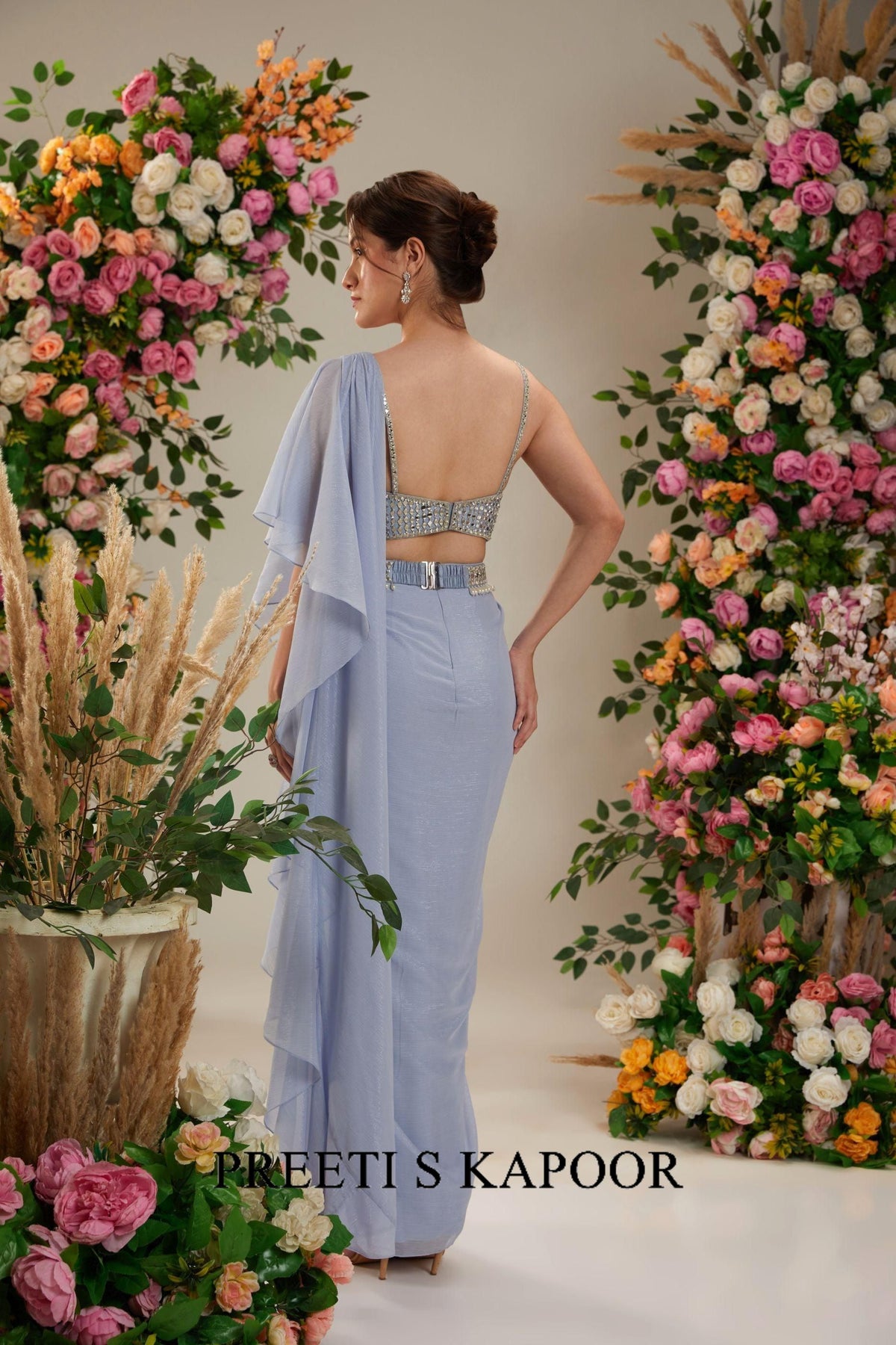 Woman in a light blue saree standing among floral decorations with 'Sreevi S Kapoor' branding.