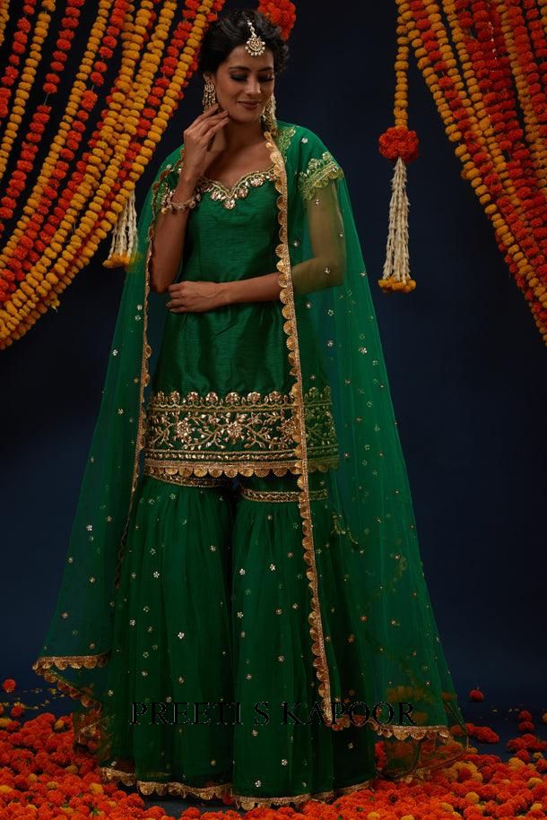 Woman in a green traditional outfit with gold embroidery against a decorated backdrop.