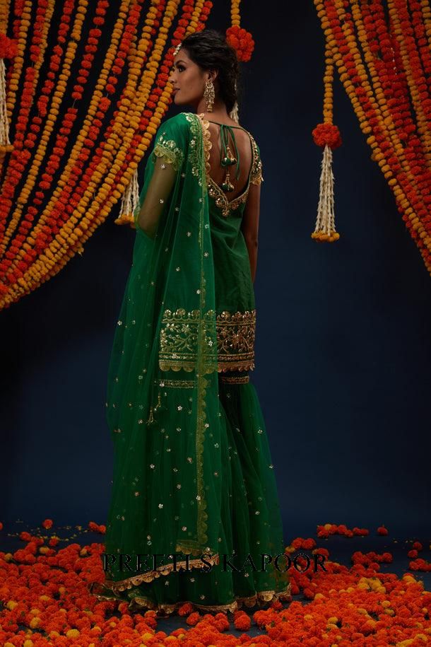 Woman in a green traditional outfit with gold embroidery standing against a decorated backdrop with orange flowers.