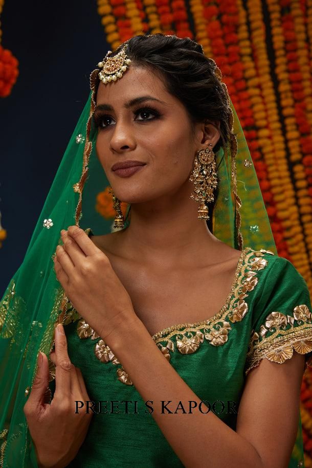 Woman in a green traditional outfit with gold embroidery, wearing a headscarf and jewelry, against a floral background.