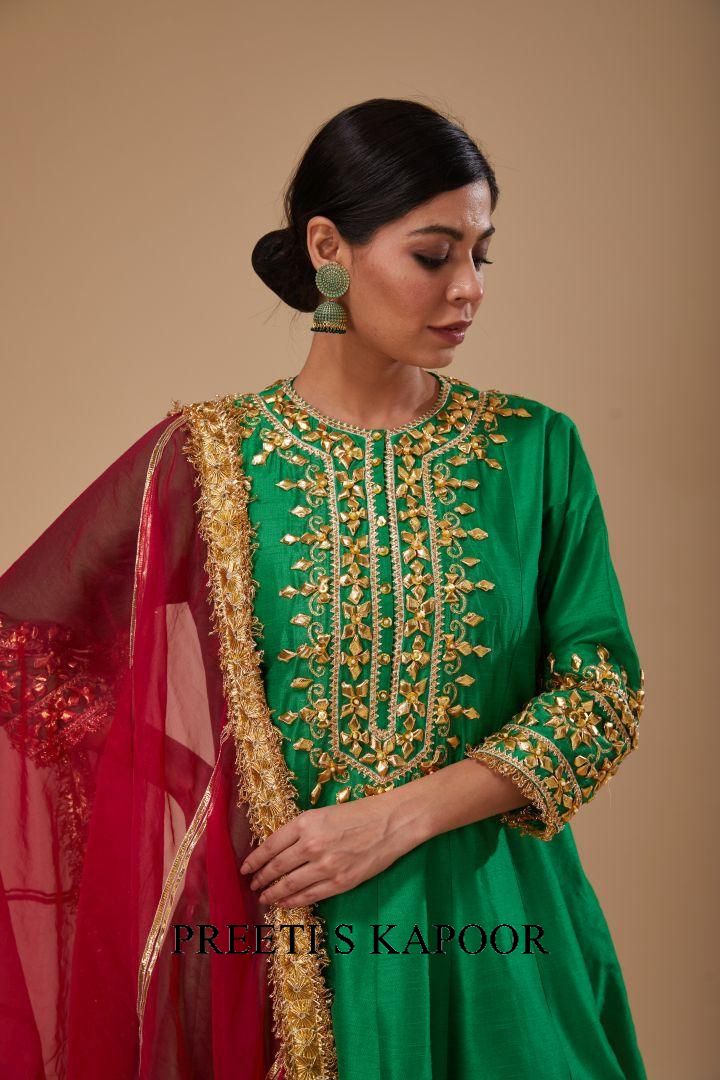Woman wearing a green embroidered outfit with gold details, holding a red dupatta, against a beige background.