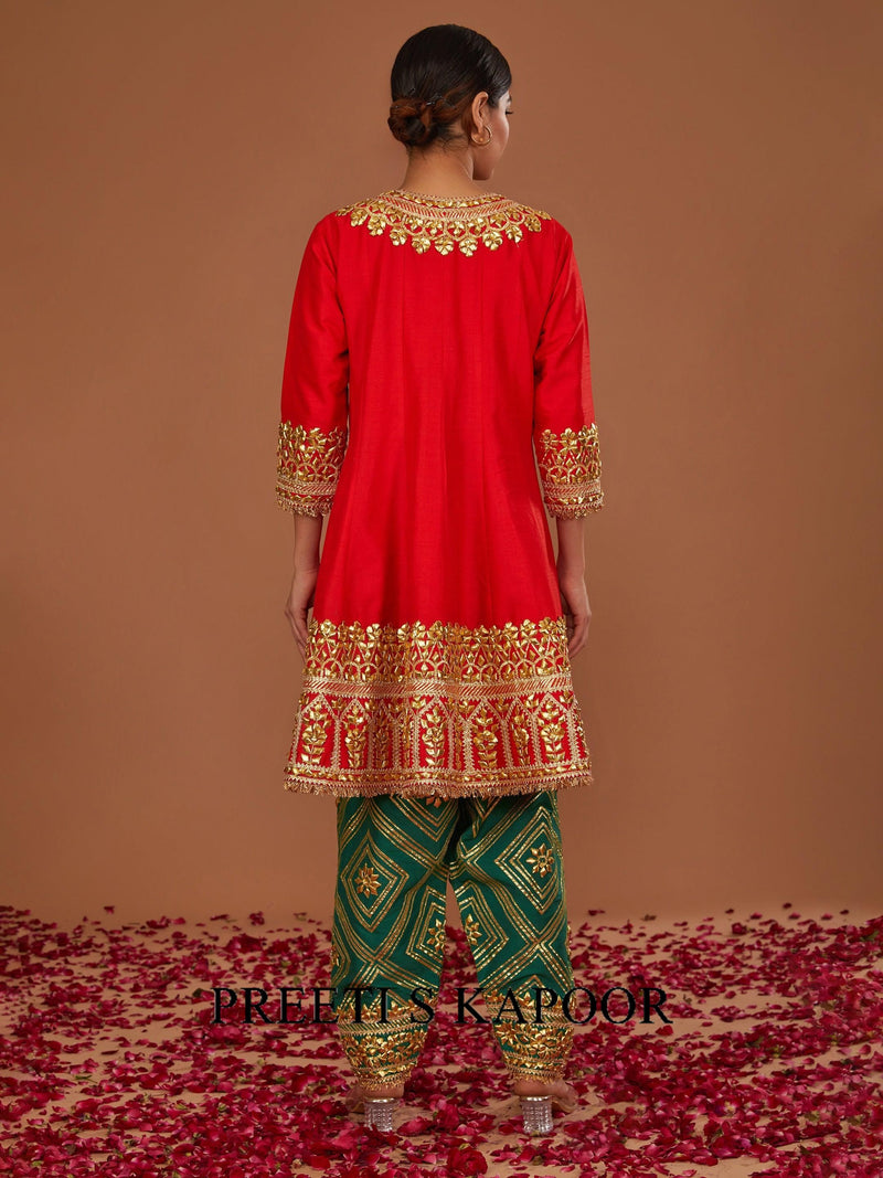 Person wearing a red and gold embroidered outfit with green pants on a brown background.