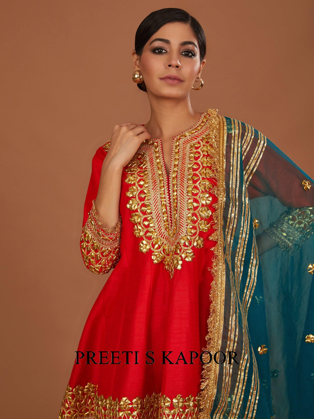Woman wearing a red and gold embroidered outfit with a blue dupatta against a brown background, branded 'Preeti S Kapoor'.