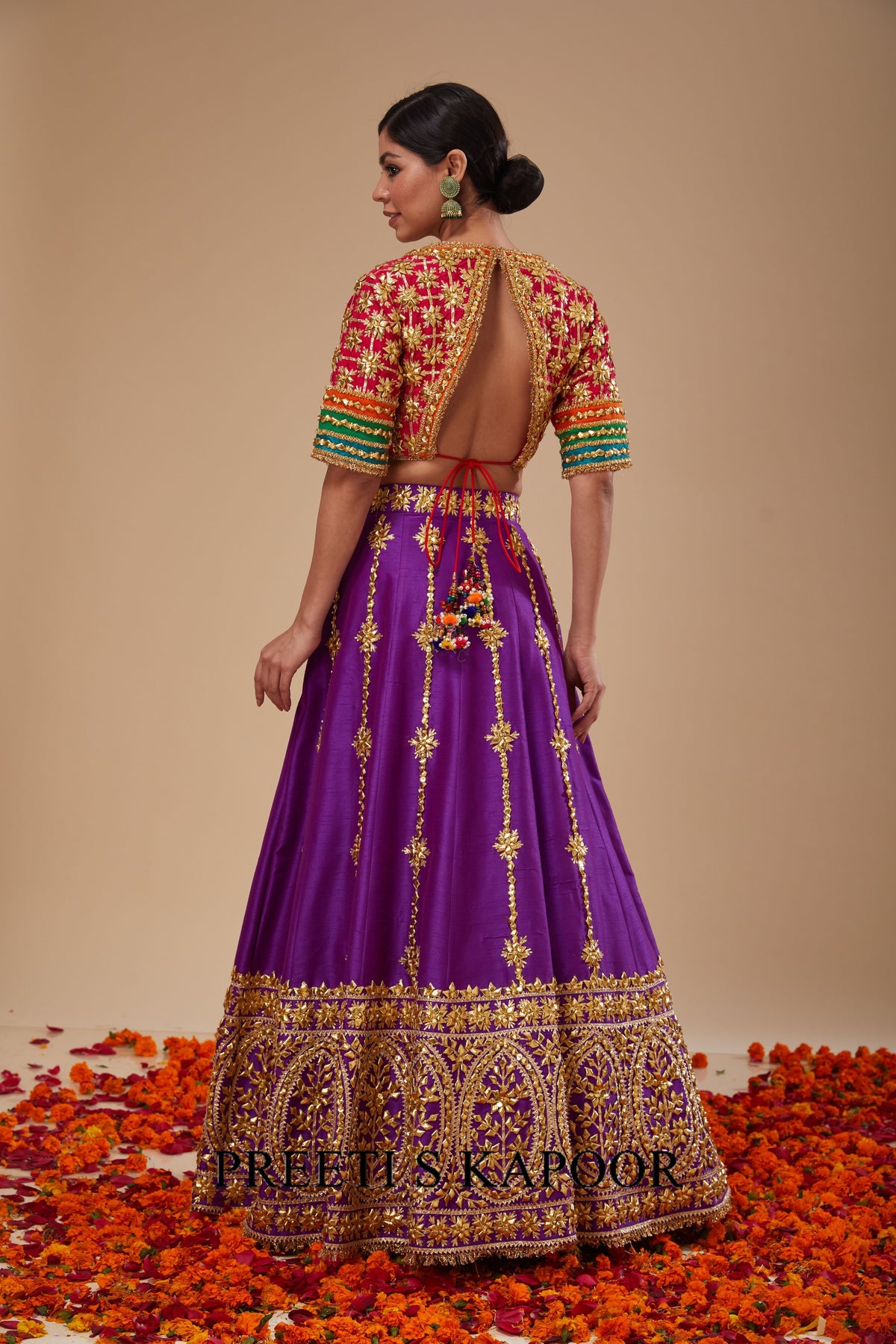 Woman wearing a traditional purple and gold outfit with a colorful blouse, standing on orange flowers against a beige background.