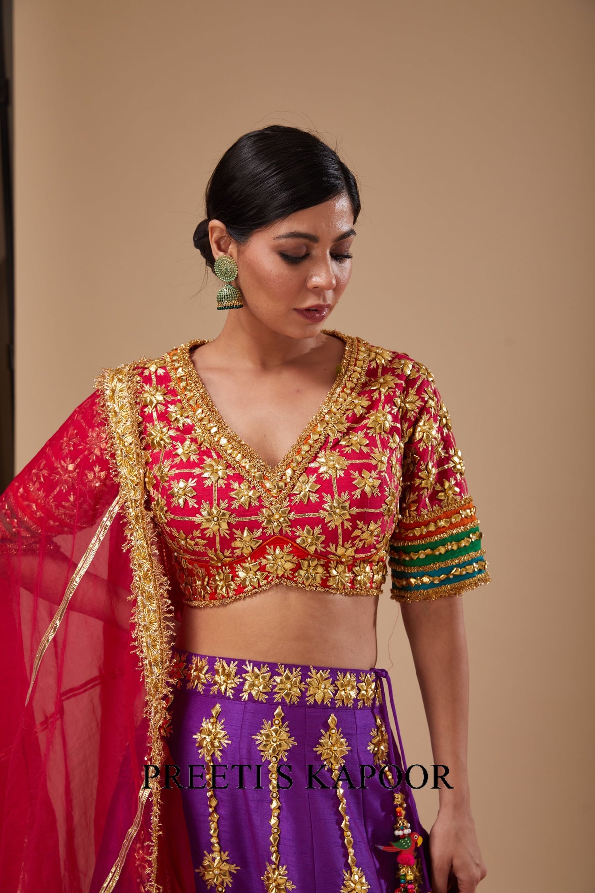 Woman wearing a traditional outfit with red and purple colors and gold embroidery, standing against a beige background.