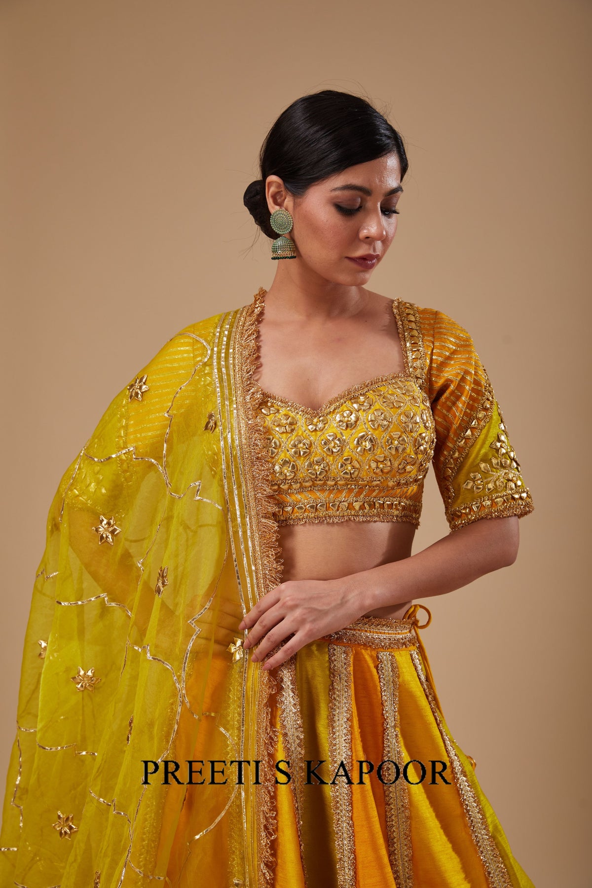 Woman wearing a yellow traditional outfit with intricate designs on a beige background, branded 'Preetis Kapoor'.
