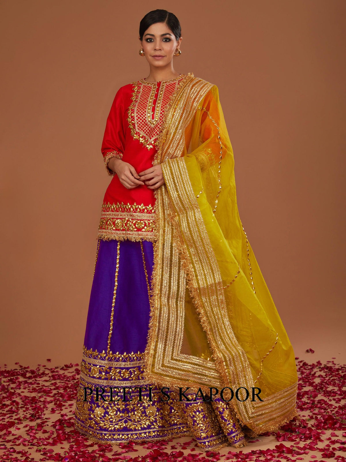 Woman wearing a traditional outfit with a red blouse, purple skirt, and yellow dupatta on a brown background.