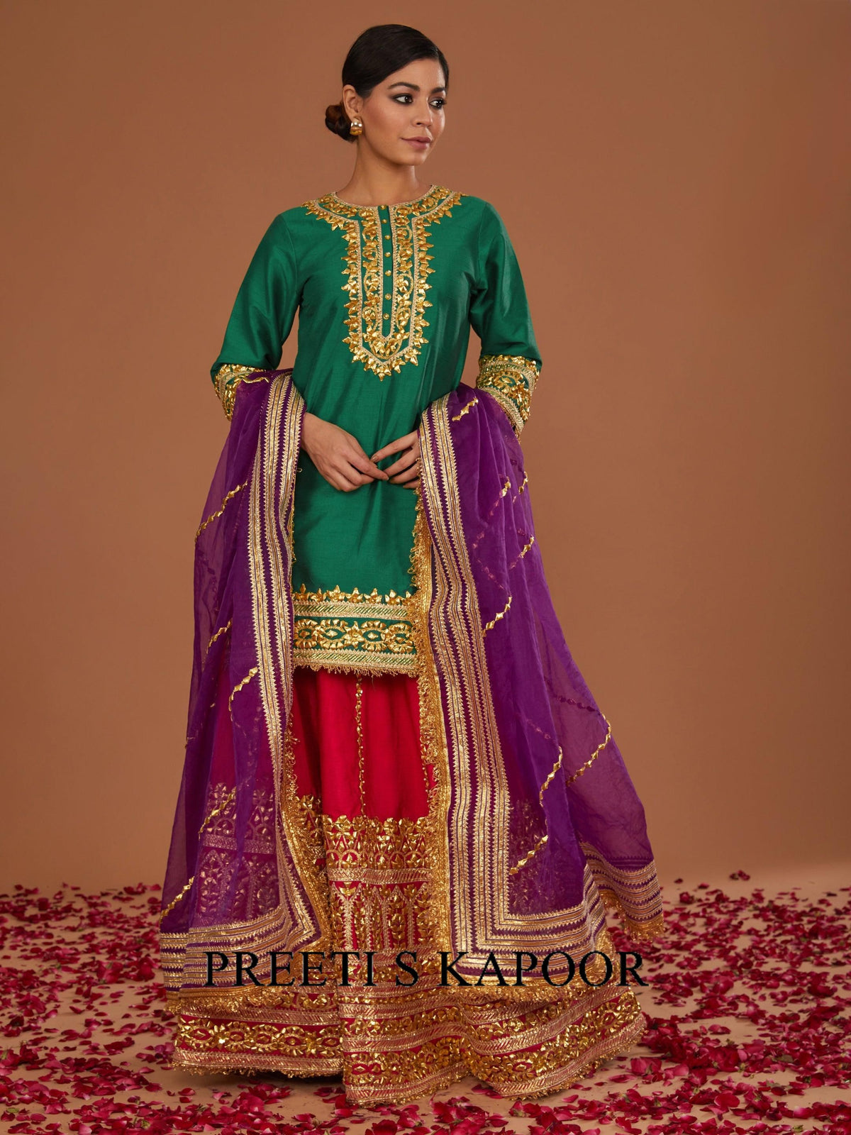 Woman wearing a traditional outfit with green top, purple dupatta, and red skirt on a brown background.