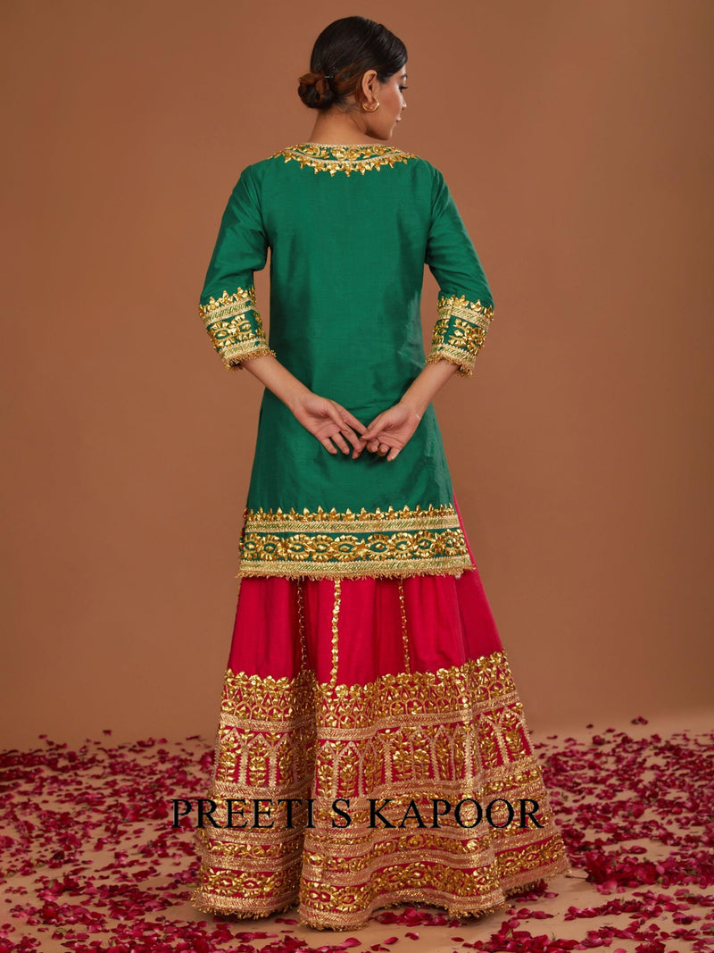 Woman wearing a green and red traditional outfit with gold embroidery on a brown background.