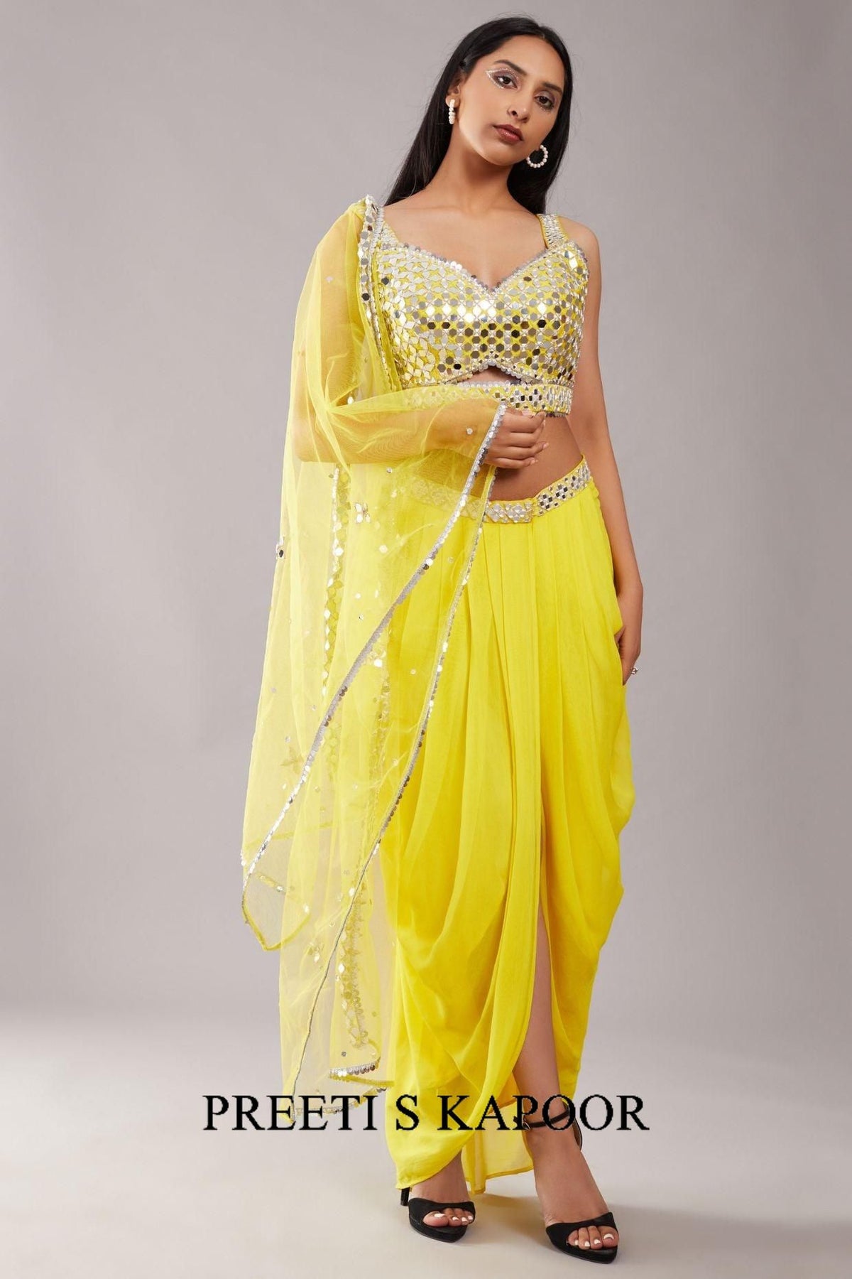 Woman wearing a yellow saree with a matching blouse, standing against a plain background. The brand 'Preeti S Kapoor' is visible.