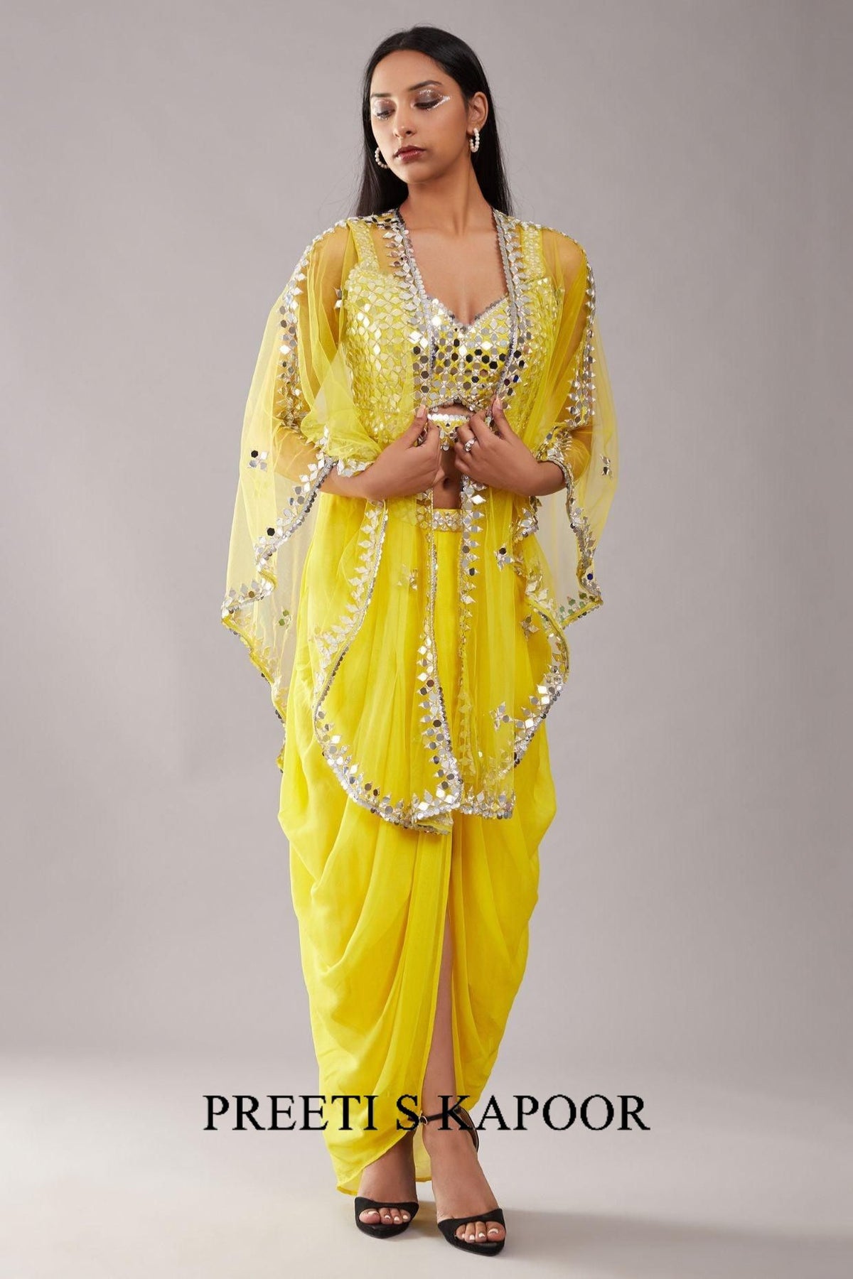 Woman wearing a yellow saree with intricate designs, holding a clutch, on a gray background. The brand 'Preeti S Kapoor' is visible.