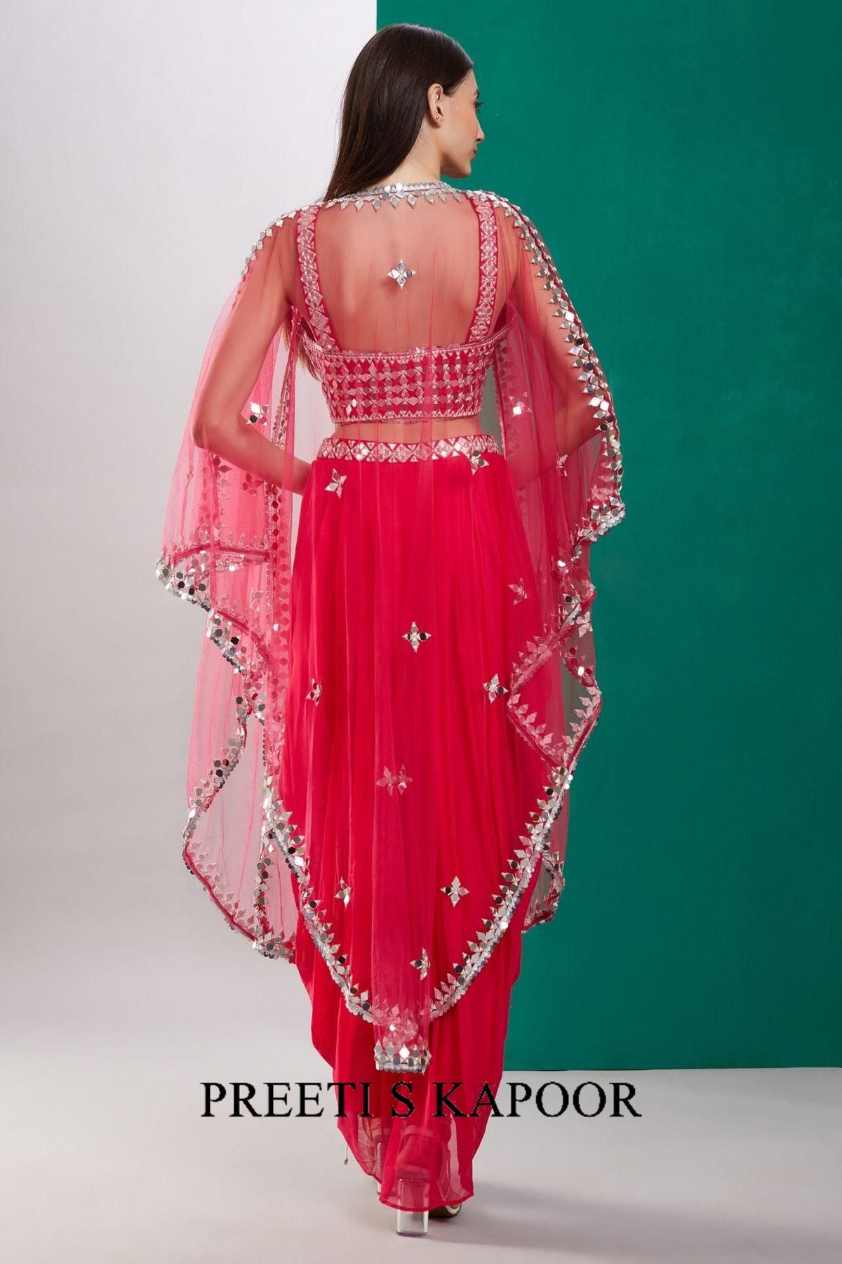 Woman wearing a pink traditional outfit with intricate designs, standing against a green and white background. Brand 'Preetus Kapoor' is visible.