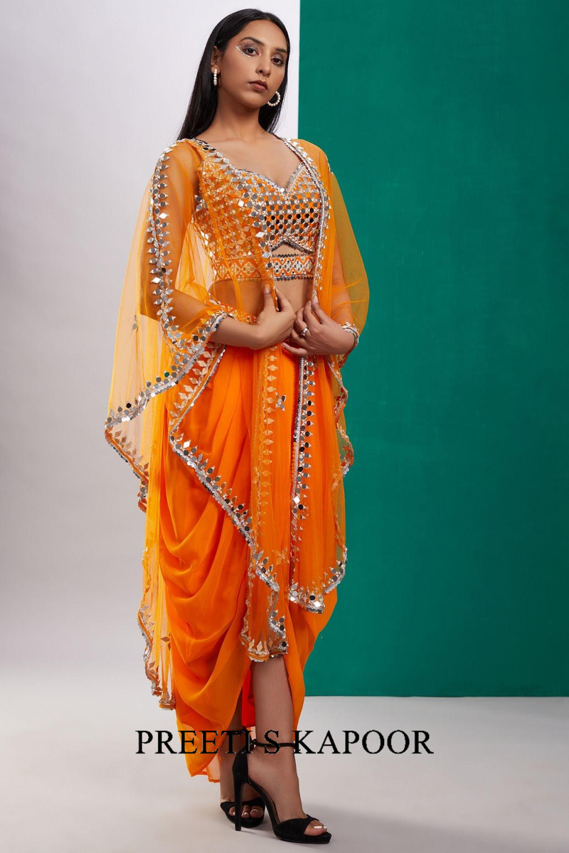 Woman wearing an orange saree with silver embellishments against a green background, with 'Preetis Kapoor' branding.