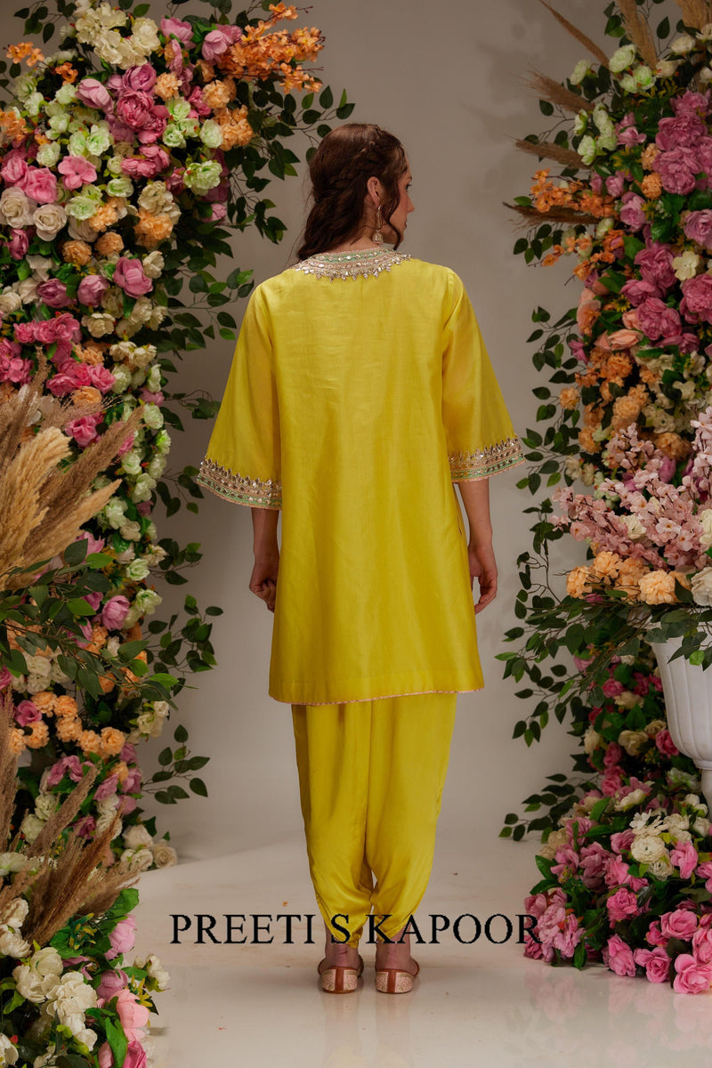 Woman in a yellow outfit standing between floral decorations with 'Preeti S. Kapoor' branding.