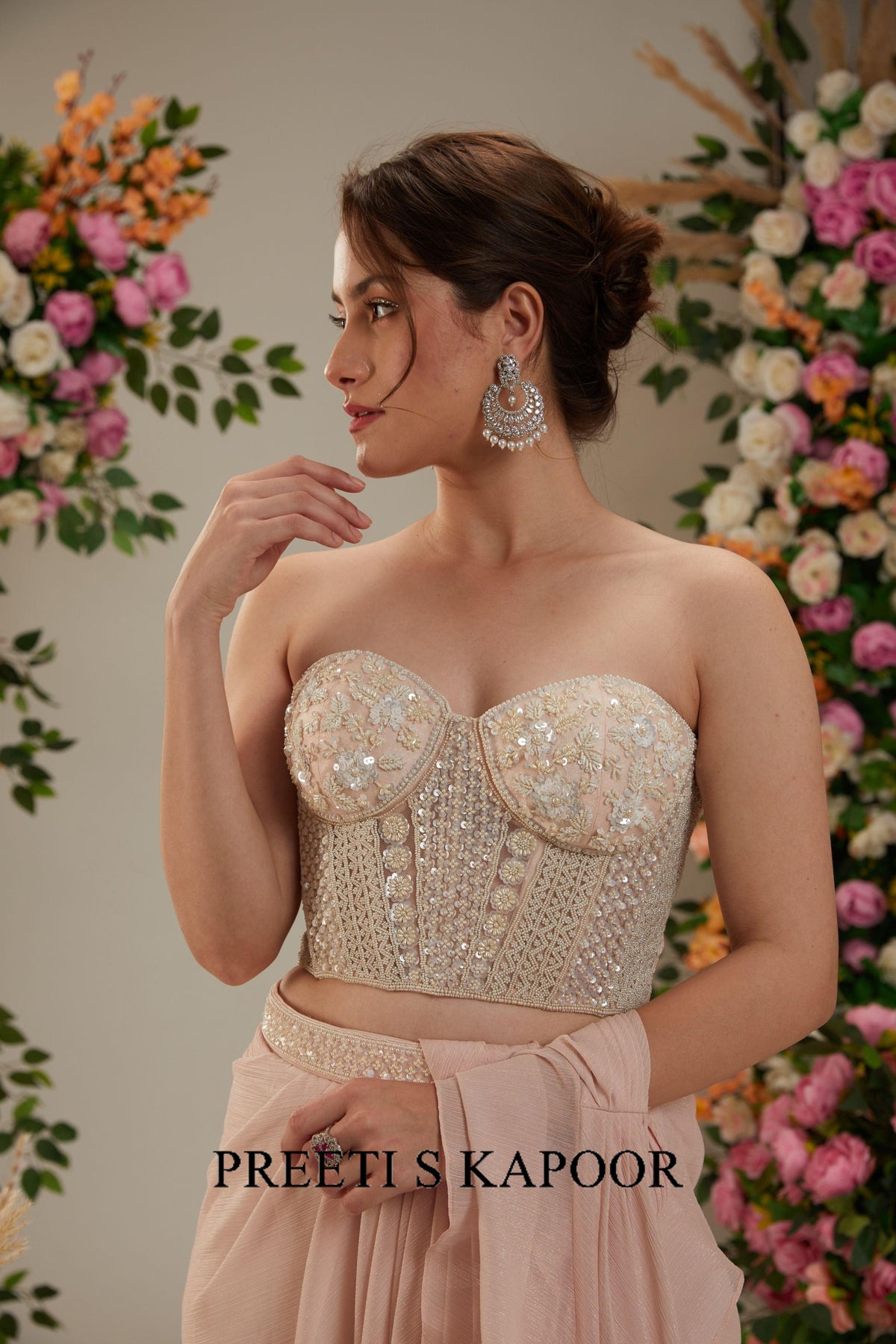 Woman wearing a beaded top and pink skirt with floral decorations in the background, branded 'Preeti S Kapoor'.