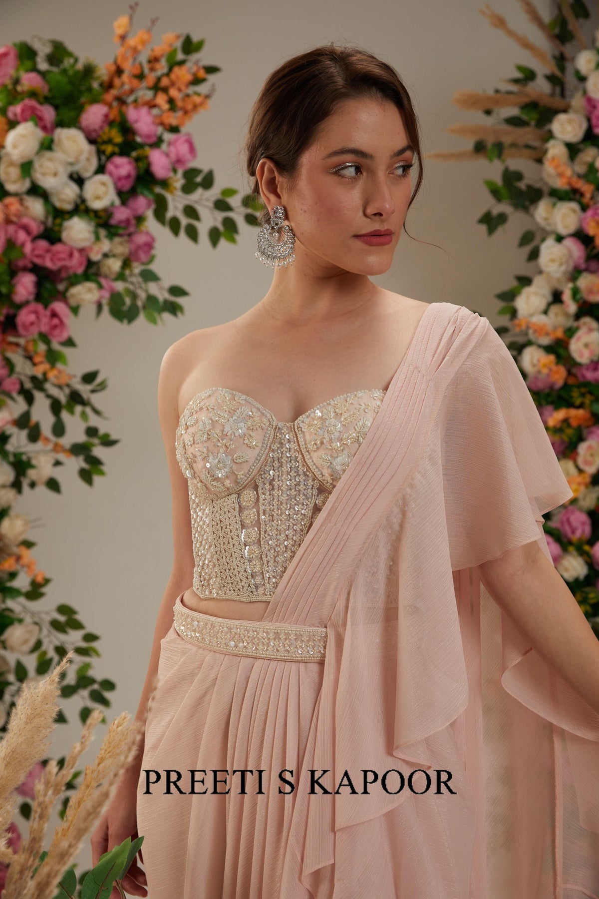 Woman in a pink saree with floral decorations in the background, featuring the brand 'Preeti S Kapoor'.