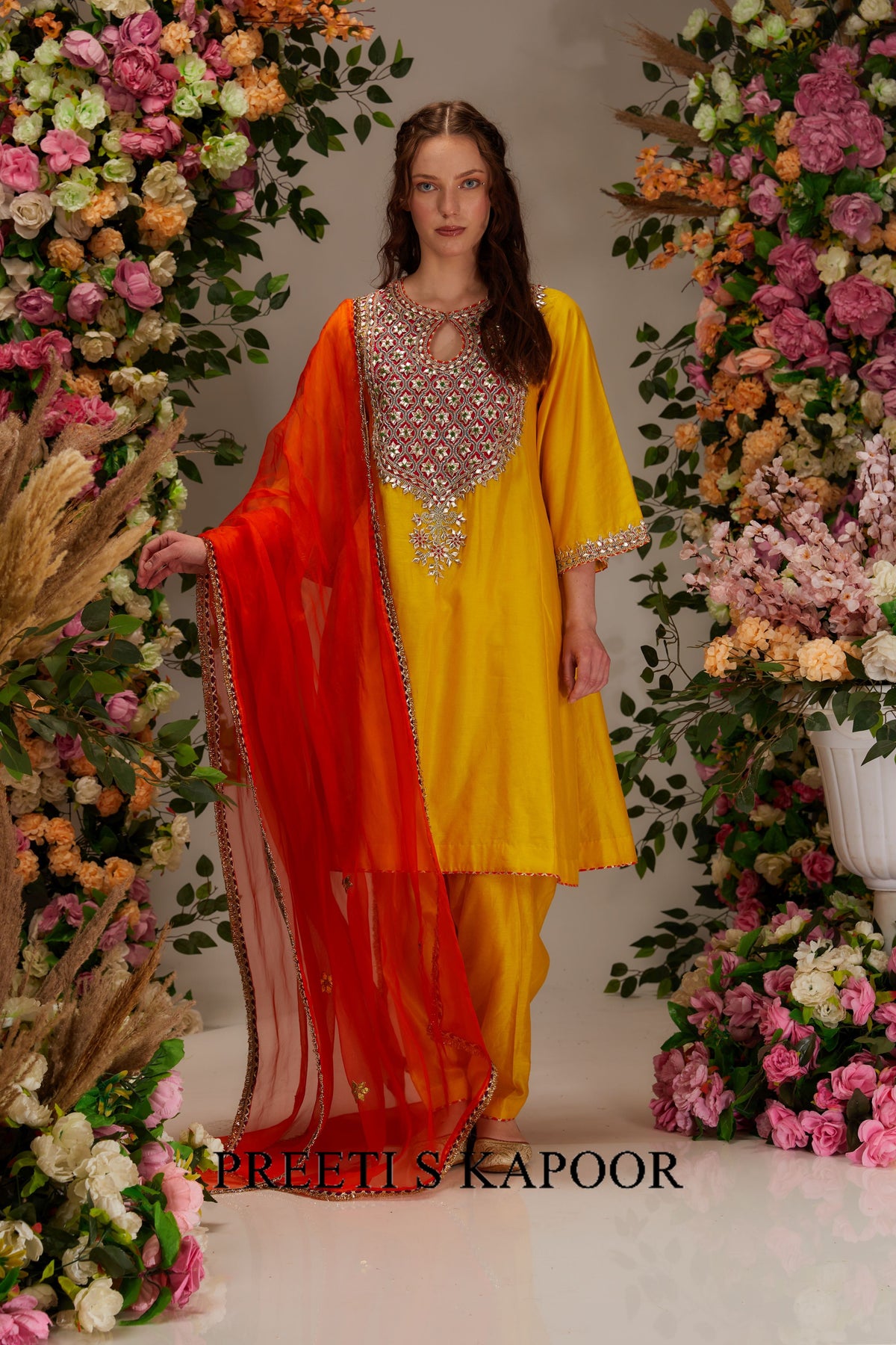 Woman in a yellow and red traditional outfit standing between floral decorations.