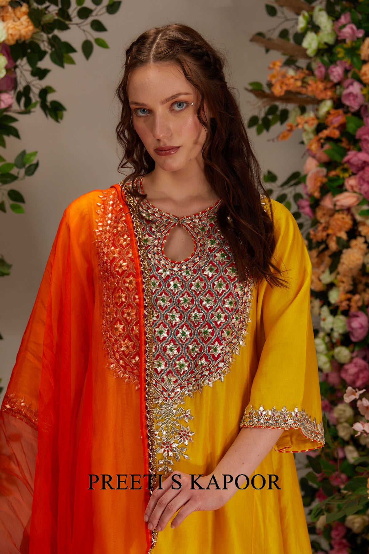Woman wearing a yellow and orange outfit with intricate patterns, standing against a floral background. Brand 'Preeti S Kapoor' visible.