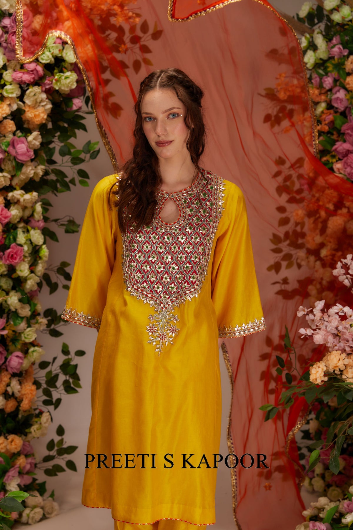 Woman in a yellow dress with intricate detailing against a floral and fabric backdrop, with 'Preeti S Kapoor' branding.