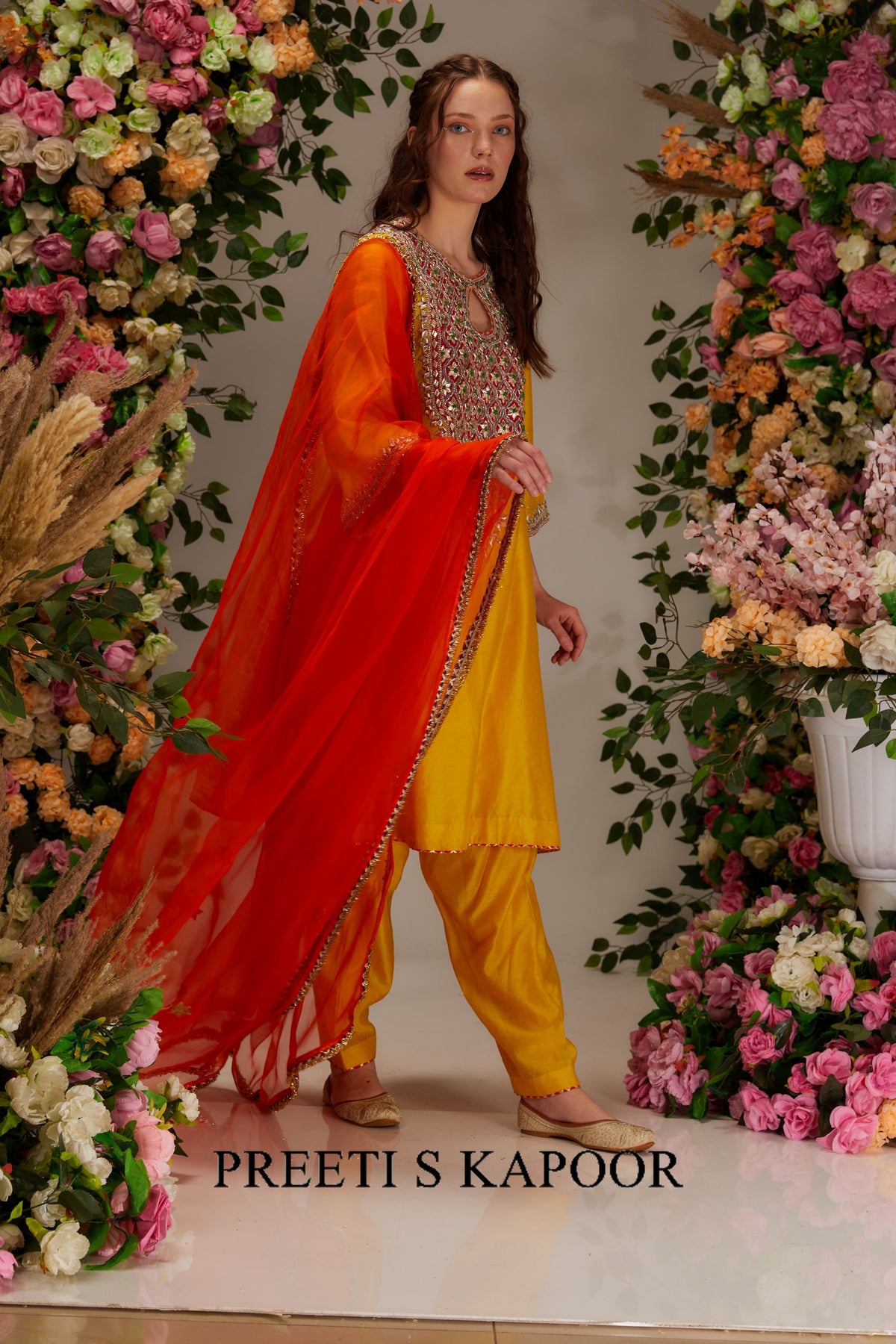 Woman in a traditional outfit with a red dupatta and yellow pants, standing in front of floral decorations.