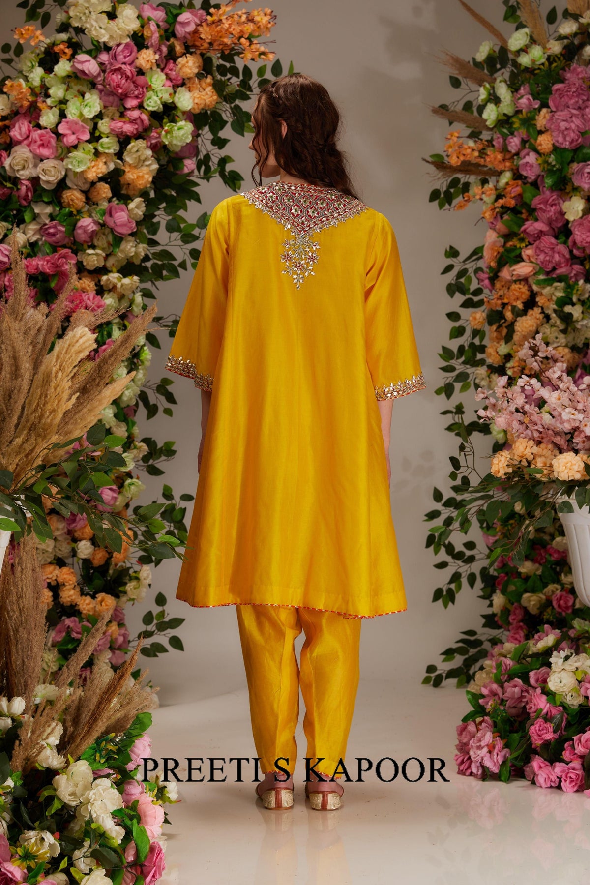 Woman in a yellow outfit standing between floral decorations with 'Preeti & Kapoor' branding.